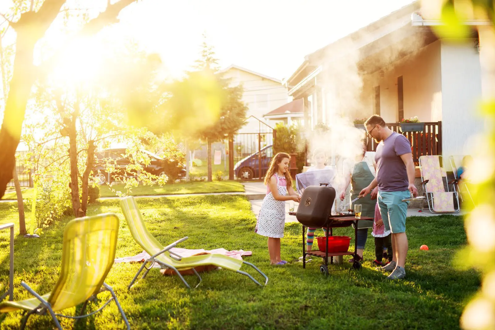 Familie grillt gemeinsam im Garten eines Ferienhauses an einem sonnigen Tag.